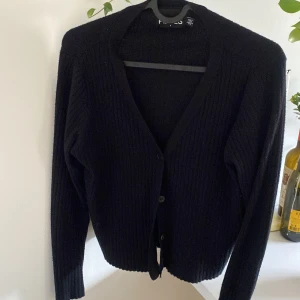 Svart ribbad kofta från Pieces - Svart ribbstickad kofta från Pieces med v-ringning och knappar framtill. Långärmad och i mjukt material som passar perfekt till lager-på-lager. Enkel och stilren design som är lätt att matcha med olika outfits.