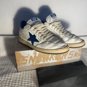 GGDB Ballstar sneakers vit/blå - Snygga GGDB Ballstar sneakers i vitt skinn med blå detaljer och stjärna på sidan. Klassisk retrostil med beige sula, perforerad tå och blå häl. Skorna har snörning och GGDB-logga på plösen. Perfekta för dig som gillar streetstyle och unika detaljer.