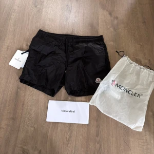 Moncler badshorts - Säljer ett par stilrena svarta badshorts från Moncler i storlek M. Mycket fint skick. Köptes på Man Of a Kind för ett par år sedan innan de köptes upp av NK. Använda ett par gånger under en sommar men sedan dess tyvärr fått hänga i garderoben.  Dustbag ingår men tyvärr inget kvitto då jag tappat bort det.  Nypris runt 3000 kr.