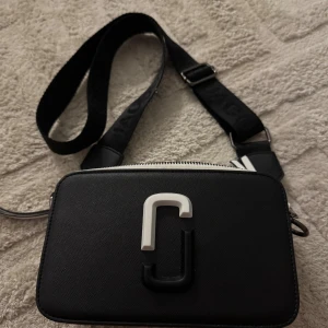 Svart och vit axelväska Marc Jacobs - Snygg axelväska från Marc Jacobs i svart och vitt med bred justerbar axelrem och dubbla dragkedjor. Väskan har en stilren rektangulär form och är dekorerad med Marc Jacobs logga framtill. Tillverkad i skinn med struktur och detaljer i silver. Väskan i sig är lite sliten som ni kan se på bild 2, men den är hel och i fint skick!