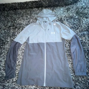 Grå vindjacka från Under Armour - Snygg tvåfärgad vindjacka från Under Armour i ljusgrått och mörkgrått. Jackan har huva, hel dragkedja framtill och diskret logga på bröstet. Perfekt för blåsiga dagar och sportiga outfits. Långärmad och med praktiska fickor.