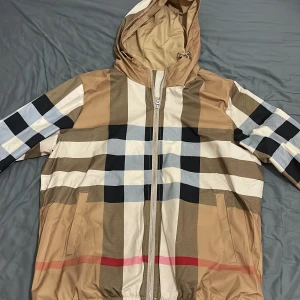 Rutig jacka med huva från Burberry - Säljer en snygg rutig jacka från Burberry i beige, svart, vit och röd. Jackan har en dragkedja framtill och en praktisk huva med dragsko. Perfekt för höst och vår! Den är i mycket bra skick och har en klassisk stil som passar till det mesta.