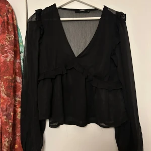 Svart omlottblus med volang från Bik Bok - Svart blus från Bik Bok i tunn, luftig polyester med omlott-skärning och v-ringning. Långa ärmar och volangdetaljer längs axlar och byst ger en romantisk vibe. Perfekt att styla med jeans eller kjol för en trendig look.