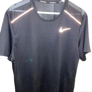 Svart Nike tränings-tshirt L - Svart t-shirt från Nike med reflekterande detaljer på axlarna och klassisk swoosh-logga på bröstet. Tillverkad i lätt och ventilerande polyester, perfekt för löpning eller gym. Rund hals och korta ärmar ger en sportig vibe.