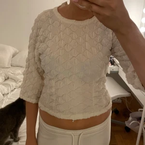 Vit quiltad topp med trekvartsärm - Supersöt vit topp med quiltad struktur och trekvartsärmar. Den har en croppad passform och rund halsringning. Perfekt för dig som gillar unika detaljer och mjuka material.