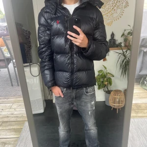 Svart pufferjacka från Polo Ralph Lauren - Säljer en svart pufferjacka från Polo Ralph Lauren med huva och dragkedja. Jackan har en glansig finish, broderad röd logga på bröstet och flera fickor med dragkedja. Perfekt för kalla dagar och riktigt snygg stil. Den har en lätt skada på ärmen som man ser på sista bilden. 