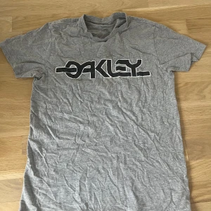 Grå Oakley t-shirt slim fit - Grå t-shirt från Oakley med stor svartvit logga över bröstet. Slim fit-modell med korta ärmar och rund hals. Tillverkad i mjuk bomull, perfekt för chill dagar eller när du vill ha en clean streetstyle-look.