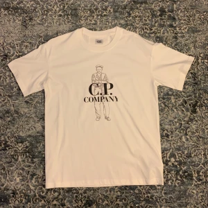 C.P. Company t-shirt - Vit t-shirt från C.P. Company med ett stilrent tryck av en figur och loggan på bröstet. Klassisk rund halsringning och korta ärmar. Tillverkad i mjuk bomull som känns skön mot huden. Perfekt för en clean och avslappnad look. Säljer då den inte passar mig tyvärr. 