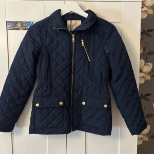 Mörkblå quiltad jacka Holly & White - Quiltad mörkblå jacka från Holly & White med guldfärgade dragkedjor och knappar. Jackan har två stora fickor framtill, en sned bröstficka med dragkedja och markerad midja. Perfekt för dig som gillar stilren och klassisk look. Står Stella på insidan av jackan.