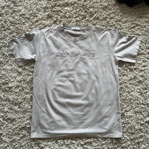 Beige t-shirt från Calvin Klein Jeans - Snygg beige t-shirt från Calvin Klein Jeans med broderad logga framtill och liten CK-detalj på ärmen. Klassisk rund hals och rak passform, perfekt för en clean och chill look. Tillverkad i mjuk bomull som känns skön mot huden.