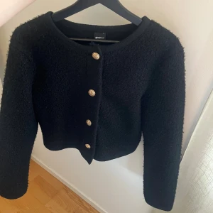 Svart kofta med guldknappar Gina Tricot - Snygg svart kofta från Gina Tricot med exklusiva guldknappar framtill. Koftan är i ett mjukt, stickat material och har lång ärm samt rund halsringning. 