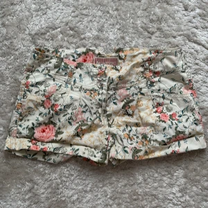 Blommiga shorts från Authentic Casuals - Snygga blommiga shorts från Authentic Casuals i ljusbeige med rosa, gröna och gula detaljer. Modellen har klassisk midja, fickor fram och bak samt uppvikta benslut. Perfekta för varma dagar och ger en chill vibe till din outfit. Midjan är medelhög men kan vikas ner om man hellre vill ha lägre! Storlek 10 men skulle säga att den passar S.  Midjemått: 39cm