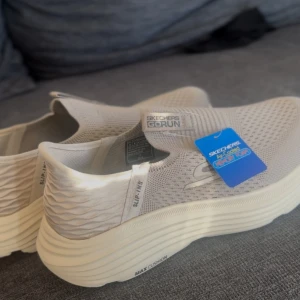 Beige Skechers GOrun slip-ins - Snygga beige Skechers GOrun slip-ins med max cushioning och Air-Cooled Memory Foam. Ovandel i stickad syntet med ventilerande mesh och elastisk passform. Platt sula och diskret S-logga på sidan. Perfekta för dig som vill ha bekväma och lätta sneakers.