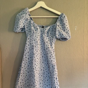 Blommig ljusblå klänning H&M Divided - Supersöt ljusblå kortklänning från H&M Divided med puffiga kortärmar och svart blommigt mönster. Klänningen har en smickrande v-ringad hals och är gjord i mjuk bomull, perfekt för varma dagar. Säljer då den inte kommer till användning längre!