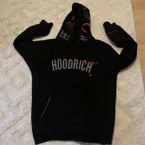 Svart HOODRICH hoodie med brodyr L - Svart hoodie från HOODRICH i storlek L med broderad logga i grått och rött på bröstet, ärmen och huvan. Hoodien har en stor huva med coola brodyrdetaljer och en klassisk känguruficka framtill. Perfekt för en streetwear-look.