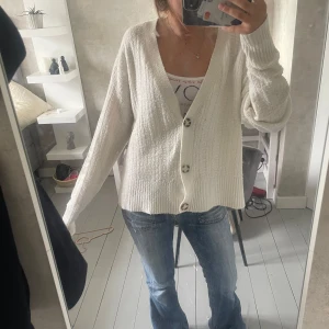 Vit kofta med knappar - Mysig vit kofta  med stora knappar framtill och v-ringad hals. Koftan har en stickad struktur och långa ärmar, perfekt att slänga på sig över ett linne eller t-shirt. Passformen är lite loose och den är superenkel att styla till jeans.
