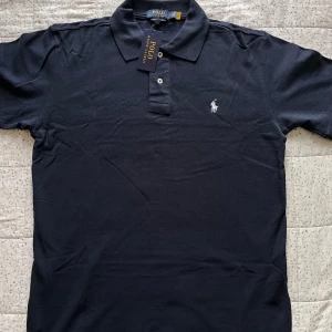 Marinblå pikétröja från Ralph Lauren - Snygg marinblå pikétröja från Polo Ralph Lauren i bomull. Klassisk krage, två knappar och ikoniska broderade loggan på bröstet. Modellen är custom slim fit och passar perfekt till jeans eller chinos. Enkel och stilren vibe.
