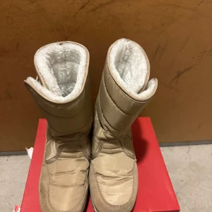 Beiga fodrade moon boots - Säljer ett par beiga moon boots med mjukt vitt foder på insidan. Skorna har quiltad ovandel och platt sula, perfekta för kalla vinterdagar. Yttermaterialet är syntet och mocka, vilket ger både värme och stil. Passar dig som vill ha varma och bekväma skor under vintern.