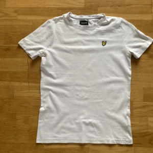 Vit Lyle & Scott t-shirt, junior 12-13år - Vit t-shirt från Lyle & Scott Junior med klassisk rund hals och korta ärmar. På bröstet sitter den ikoniska broderade gula örnen. T-shirten är gjord i mjuk bomull och har en clean, stilren look som funkar till jeans eller shorts.