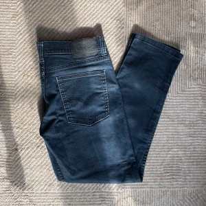 Levi's 517 blå jeans - Säljer ett par Levi's 517 jeans i snygg mörkblå tvätt med klassisk bootcut-passform. Jeansen har fem fickor, tydliga sömmar och ett läderpatch bak i midjan. Materialet är slitstarkt denim som sitter skönt och ger en cool look. Storlek 32/32. 