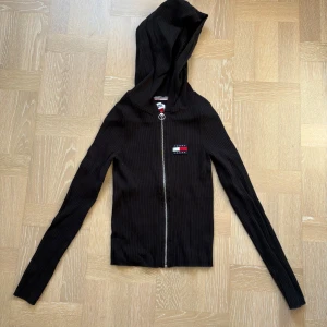 Svart ribbad hoodie - Svart ribbad hoodie från Tommy Jeans med dragkedja och logga på bröstet. Långärmad med huva och ringformad dragkedjedetalj.