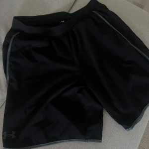 Svarta träningsshorts från Under Armour - Svarta shorts från Under Armour med grå detaljer längs sidorna. Perfekta för träning, med elastisk midja och lätt, syntetiskt material som andas. Diskret logga nere på vänster ben. Snygg och sportig look för gymmet eller löprundan.