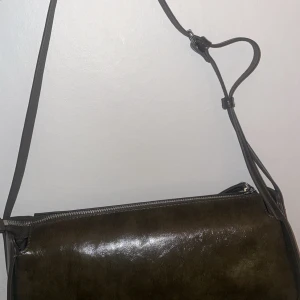 Olivgrön axelväska i skinn - Snygg olivgrön axelväska i skinn med stilren, avlång form och justerbar axelrem. Väskan har en enkel design med dragkedja upptill och passar perfekt för dig som gillar minimalistiska accessoarer.