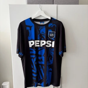Pepsi Max fotbollströja  - Säljer en svart och blå fotbollströja från Pepsi i storlek M. Tröjan har korta ärmar, rund hals och ett grafiskt mönster med blå detaljer och Pepsi-logga på bröstet. Materialet är lätt och andas, perfekt för träning eller match. Snygg och unik design med stora vita Pepsi-trycket framtill.