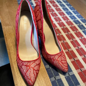 Röda spetsiga pumps från Moda Aire - Snygga röda pumps från Moda Aire med spetsig tå och hög klack. Skorna har ett unikt mönster i röd spets över en ljus bas och röda detaljer längs kanterna. Perfekta för dig som vill sticka ut med en färgstark och elegant look.