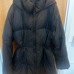 Svart lång pufferjacka med huva - Säljer en svart pufferjacka med stor, mysig huva och dragkedja framtill. Jackan har snörning i midjan för att justera passformen och två praktiska fickor. Perfekt för kalla dagar när du vill hålla dig varm och ändå se chill ut.