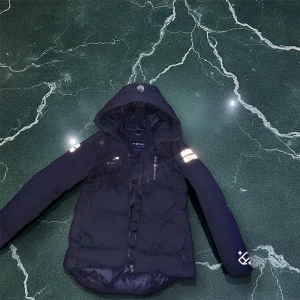 Svart dunjacka med huva från sail racing  - Svart dunjacka från sail racing  med huva och reflekterande detaljer på ärmarna. Jackan har dragkedja framtill, flera fickor och en sportig look. Perfekt för kalla dagar när du vill hålla värmen och ändå se snygg ut. Blir perfekt nu till vintern 