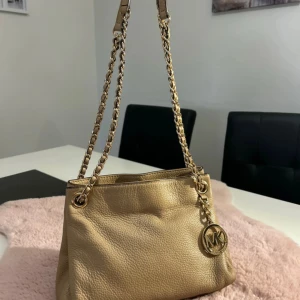 Beige axelväska från Michael Kors - Snygg beige axelväska från Michael Kors i äkta skinn med kedjedetaljer i guld och klassisk MK-logga. Väskan har en mjuk, kornig struktur och stilren form som passar perfekt till din vardagslook. Lång axelrem i kedja för extra statement.