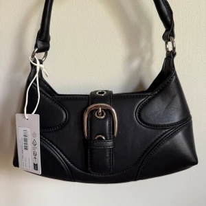 Svart axelväska med spänne från NA-KD - Snygg liten svart axelväska från NA-KD. Väskan har ett coolt silverfärgat spänne framtill och bred axelrem. Stilren design med markerade sömmar och detaljer som ger en trendig vibe. Perfekt storlek för det viktigaste.