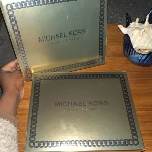 Michael Kors Pour Femme presentbox - Lyxig presentbox från Michael Kors Pour Femme med en parfymflaska i guld och svart design, samt matchande duschgel och body lotion i nude-färgade tuber. Allt levereras i en snygg guldfärgad ask med kedjemönster runt kanten. Perfekt för dig som älskar eleganta dofter och exklusiv känsla.