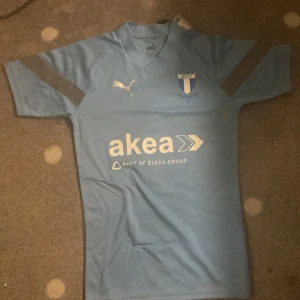 Malmö FF ljusblå matchtröja Puma - Snygg ljusblå fotbollströja från Puma med Malmö FF-logga på bröstet och sponsortryck från akea. Tröjan har korta ärmar med gråa detaljer och är gjord i ett lätt, ventilerande material som passar perfekt på planen. Klassisk V-ringning och modern passform. MEDIUM STORLEK