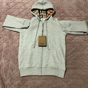 Grå hoodie från Burberry med dragkedja - Snygg grå hoodie från Burberry med klassiskt rutigt foder i huvan och broderad logga på bröstet. Tröjan har dragkedja framtill, snörning i huvan och ribbade muddar. Perfekt för en chill och stilren look.