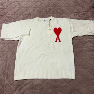 Vit stickad tröja från Ami Paris - Vit stickad tröja från Ami Paris med röd hjärt- och A-logga på bröstet. Tröjan har rund halsringning, ribbade muddar och är långärmad. Perfekt för dig som gillar stilrena plagg med en twist.