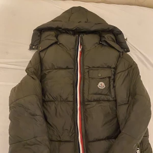 Grön Moncler dunjacka med huva - Säljer en grön Moncler dunjacka med avtagbar huva och coolt Moncler-märke på bröstet. Jackan har en ficka med knapp framtill, dragkedja med röd-vita detaljer och ser riktigt fet ut till vintern. Insidan har en unik serietidningsetikett. Perfekt för dig som vill sticka ut.