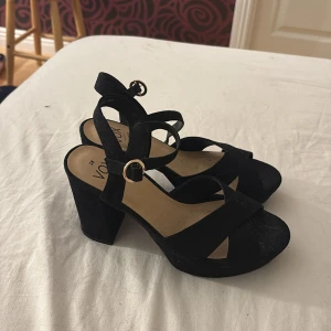 Svarta sandaler med klack från Vox - Snygga svarta sandaler från Vox med bred klack och öppen tå. Skorna har korsade remmar framtill och justerbart spänne i guld vid ankeln. Tillverkade i veganskt material med en plattformsula som ger extra höjd och stabilitet.