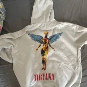 Vit Nirvana hoodie med tryck - Säljer en vit hoodie med stort Nirvana-tryck och ikonisk ängelgrafik både fram och bak. Hoodien har huva med snörning och en stor magficka. Perfekt för dig som gillar bandmerch och vill sticka ut med en klassisk look.