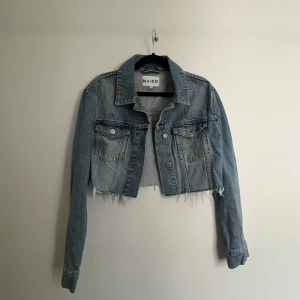 Croppad jeansjacka från NA-KD - Snygg croppad jeansjacka från NA-KD i ljusblå denim. Jackan har rå, fransig nederkant, klassisk krage och två bröstfickor med knappar. Perfekt för dig som vill ha en trendig och avslappnad look.