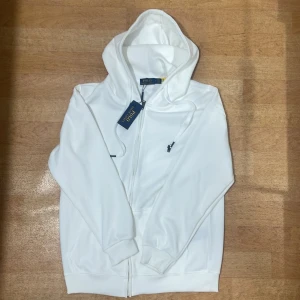 Vit hoodie från Polo Ralph Lauren - Snygg vit hoodie från Polo Ralph Lauren med klassisk logga broderad i svart på bröstet. Tröjan har huva med snörning, dragkedja framtill och ribbade muddar vid ärmar och nederkant. Perfekt för dig som gillar stilren och enkel design.