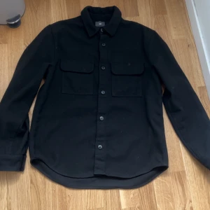 Svart overshirt från H&M - Stilren svart overshirt från H&M med klassisk krage, två stora bröstfickor med lock och knäppning framtill. Skjortan har lång ärm och är tillverkad i ett mjukt material som passar perfekt för lager-på-lager-stil.
