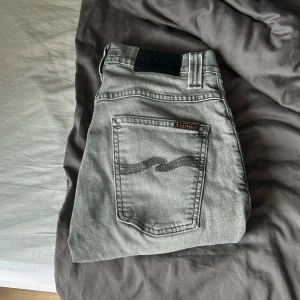 Grå jeans från Nudie Jeans, slim fit - Säljer ett par grå jeans från Nudie Jeans i modellen slim fit. De har klassisk femficksdesign, svarta detaljer på bakfickan och snyggt slitna partier. Jeansen är raka i benen och har normal passform, perfekta för dig som gillar stilrena och moderna jeans. Jeansen har dock ett pytte litet hål som man ser på sista bilden och dom är uppsydda till l28 modell grim tim 