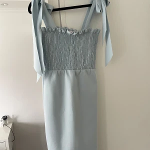 Ljusblå långklänning med knytband - Supersöt ljusblå långklänning med smockad överdel och breda axelband som knyts i stora rosetter på axlarna. Klänningen har en rak siluett och är ärmlös, perfekt för sommarens alla tillfällen. Materialet känns mjukt och lätt mot huden.