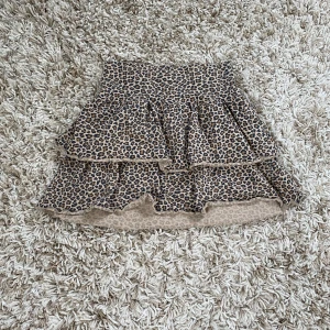 Leopardmönstrad volangkjol från H&M - Säljer en kort kjol från H&M med leopardmönster i beige och svart. Kjolen har två lager volanger som ger en snygg och lekfull look. Materialet är mjukt och stretchigt, perfekt för dig som gillar att sticka ut med coola detaljer.