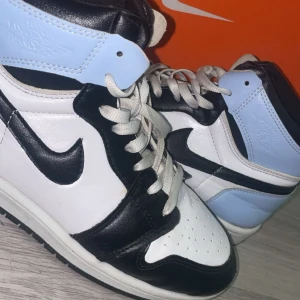 Nike Air Jordan 1 svart/vit/blå - Snygga Nike Air Jordan 1 sneakers i svart, vitt och ljusblått skinn. Klassisk high-top modell med svart swoosh och vita skosnören. Perfekta för dig som vill sticka ut med en ikonisk streetwear-look.