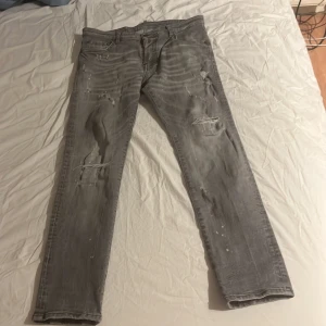 Grå Dsquared2 jeans med slitningar - Säljer ett par grå jeans från Dsquared2 med coola slitningar och färgstänk. Jeansen har rak passform, klassiska fem fickor och snygga detaljer som ger en trendig look. Perfekta för dig som gillar orten- stil och vill sticka ut lite extra. Pris ej hugget i sten.