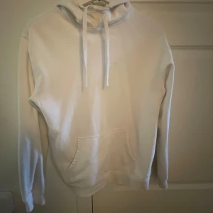 Vit hoodie med huva och ficka - En stilren vit hoodie med huva och snörning, samt en stor magficka framtill. Tröjan har lång ärm och är tillverkad i mjukt material, perfekt för en avslappnad look. 