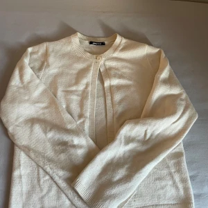 Krämvit kofta från Gina Tricot - Superfin krämvit kofta från Gina Tricot med rund halsringning och en knapp upptill. Koftan är långärmad och har ribbade kanter vid ärmslut och nederkant. Perfekt att slänga över ett linne för en chill look.